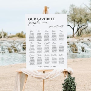May include: A white seating chart on a wooden easel with the text "OUR FAVORITE PEOPLE" in a script font. The chart lists names under numbers one through fifteen. The names Alyssa & Joseph and a date are at the bottom. Beige fabric and greenery decorate the easel.