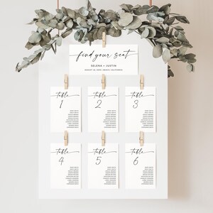 May include: A white seating chart with the text "find your seat" and the names "Selena + Justin" printed on it. The chart is hung with string and decorated with eucalyptus leaves. Six cards with table numbers and guest names are clipped to the chart.