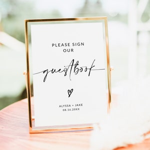 May include: A gold-framed sign with white background and black text that reads "Please sign our guestbook" with a heart symbol and the names "Alyssa + Jake" and the date "08.16.20XX".