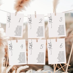 May include: Six white cards with black calligraphy that reads "Table" and a number from 1 to 6. Each card lists a set of names, including Mary Smith, Emily Smith, Ashley Brown, Joseph Brown, Megan Wilson, Kathleen Miller, Charles Johnson, Linda Johnson, George Davis, and Stephanie Davis.
