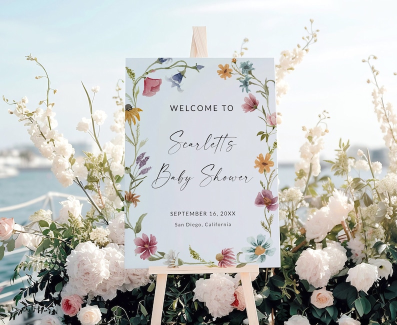 Pu&ograve; includere: Un cartello rettangolare bianco su un cavalletto di legno con un bordo di fiori ad acquerello. Il testo recita: WELCOME TO, Scarlett's Baby Shower, SEPTEMBER 16, 20XX, San Diego, California. Il design include vari fiori di campo.