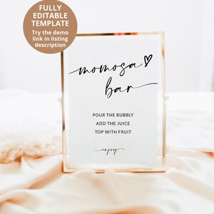 May include: A gold-framed sign with the text "mimosa bar" in black cursive script with a heart symbol. The sign also includes instructions for making mimosas: "Pour the bubbly, add the juice, top with fruit, enjoy."