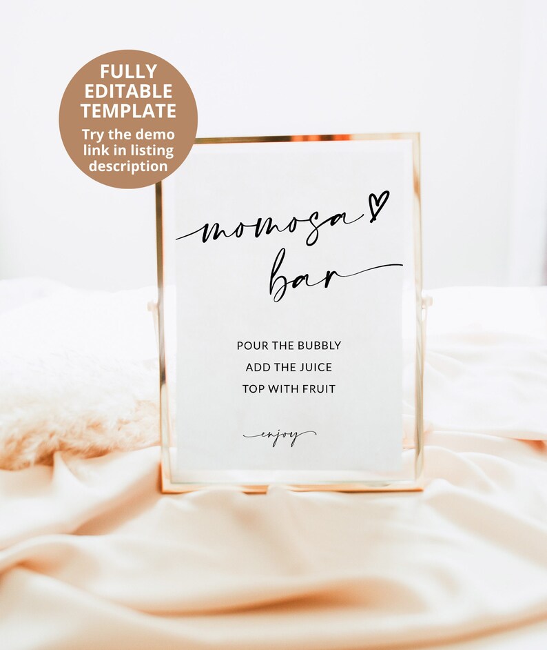 May include: A gold-framed sign with the text "mimosa bar" in black cursive script with a heart symbol. The sign also includes instructions for making mimosas: "Pour the bubbly, add the juice, top with fruit, enjoy."