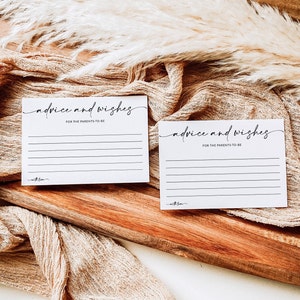 Puede incluir: Dos tarjetas blancas con texto negro que dice "consejos y deseos para los futuros padres". Las tarjetas est&aacute;n sobre una superficie de madera con una tela beige drapeada sobre ella.