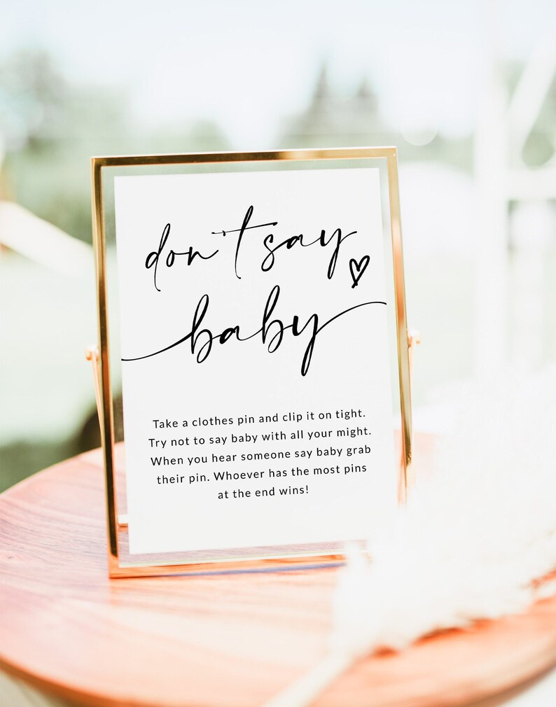 May include: A white sign in a gold frame with the text "don't say baby" in black cursive writing. The sign also includes instructions for a game: "Take a clothes pin and clip it on tight. Try not to say baby with all your might. When you hear someone say baby grab their pin. Whoever has the most pins at the end wins!"