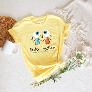 May include: Yellow t-shirt featuring a graphic of two cartoon eyes holding hands. The text "Better Together" and "#VisionTherapy" are printed below the graphic.