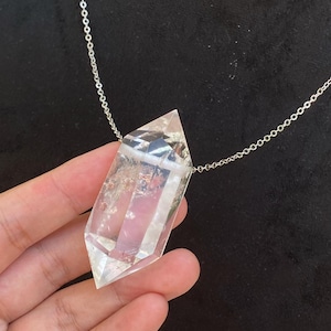 Puede incluir: Un colgante de cristal de cuarzo transparente de doble terminación suspendido de una fina cadena de metal plateado. El cristal presenta inclusiones naturales y facetas que captan la luz, creando una forma geométrica hexagonal alargada.