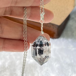Puede incluir: Un colgante de cristal transparente, facetado y de doble terminación con inclusiones negras internas, suspendido de una fina cadena de metal plateado. El cristal está asegurado en la parte superior con un alambre plateado y una anilla.