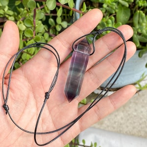 May include: A purple and green fluorite crystal pendant with a black cord. The crystal is a long, hexagonal shape with a pointed tip. The cord is knotted at the top to form a loop for wearing. The background is blurred with green foliage.