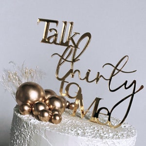 May include: A white cake with gold and black decorations. The cake is decorated with gold spheres, a black line drawing of a face, and a gold topper that says "Talk Thirty To Me".