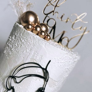 May include: White cake with gold and black decorations. The cake is decorated with a black wire sculpture of a face, gold balls, and a gold topper that says "Talk Thirty To Me".