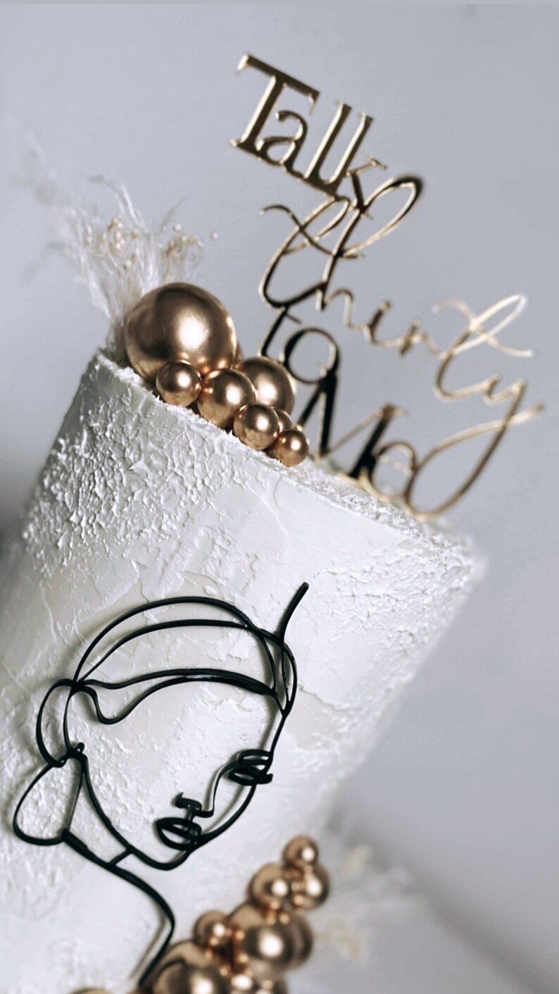 May include: White cake with gold and black decorations. The cake is decorated with a black wire sculpture of a face, gold balls, and a gold topper that says "Talk Thirty To Me".