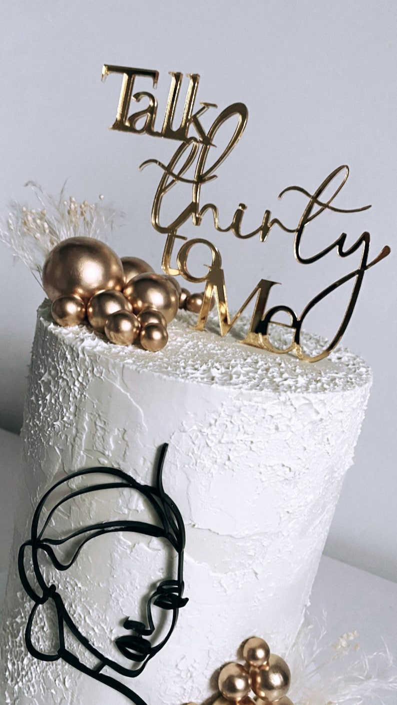May include: A white cake with gold and black decorations. The cake is decorated with gold spheres, a black line drawing of a face, and a gold topper that says "Talk Thirty To Me".