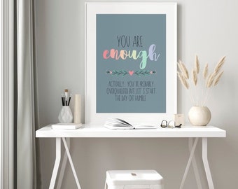 May include: A framed print with the words "YOU ARE enough" in colorful script. Below the main text is the phrase "ACTUALLY, YOU'RE PROBABLY OVERQUALIFIED BUT LET'S START THE DAY OFF HUMBLE". The print is on a white desk with a white stool.