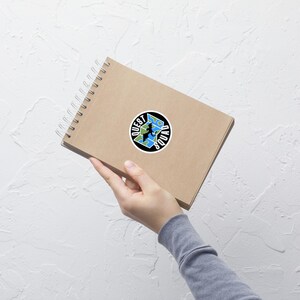 May include: A brown notebook with a spiral binding and a black and white sticker on the cover. The sticker features a globe with a silhouette of a person standing on top of it. The text "QUEST SQUAD" is written around the globe.