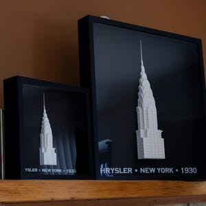 May include: Two framed art pieces featuring a white 3D model of the Chrysler Building against a black background. The text "CHRYSLER • NEW YORK • 1930" and "YSLER • NEW YORK • 1930" is visible. The frames are black and sit on a wooden shelf.