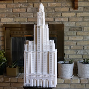 May include: A white 3D printed model of a tall building with a clock tower. The model is sitting on a table in front of a brick fireplace.