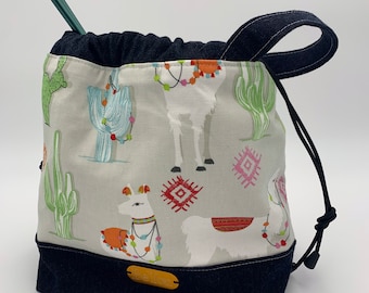May include: A drawstring bag with a denim handle and bottom. The bag is made of a white fabric with a pattern of llamas wearing colorful string lights and cacti. The bag has a brown leather tag with the word "Llama" on it.