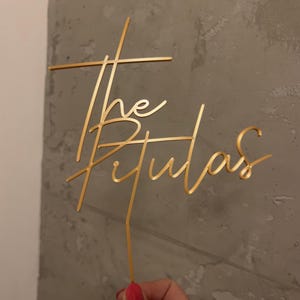 May include: A gold-colored cake topper featuring the text "The Tulas" in a cursive script font. The design includes a vertical stem at the bottom for insertion into a cake, with the lettering integrated into a cross-like structure.