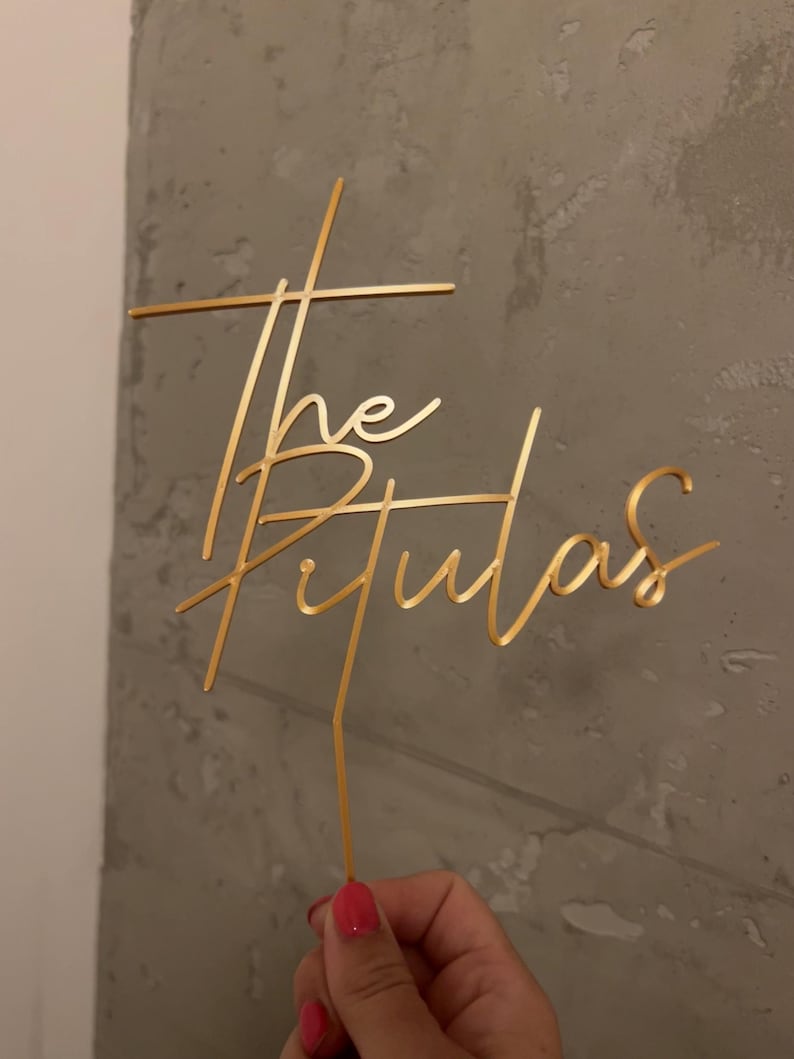 May include: A gold-colored cake topper featuring the text "The Tulas" in a cursive script font. The design includes a vertical stem at the bottom for insertion into a cake, with the lettering integrated into a cross-like structure.