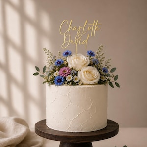 May include: A white frosted cake with a textured finish, topped with a floral arrangement of white roses, blue cornflowers, and small pink and white daisies. A gold-colored cake topper displays the names Charlotte & David in script.