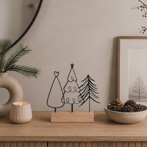 Könnte beinhalten: Ein dekoratives Tischornament mit drei stilisierten schwarzen Metallbäumen auf einem rechteckigen Sockel aus hellem Holz. Die Bäume bestehen aus einer einfachen Dreiecksform mit Herzspitze, einem geschwungenen Liniendesign mit Stern und einer Tanne.
