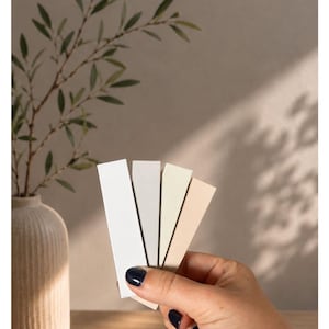 May include: A set of four rectangular color swatches in shades of white, muted white, sand, and beige. The text "Unsere Lights" and "WEIB - MUTED WHITE - SAND - BEIGE" is printed above the swatches, which are held in a fan-like arrangement.