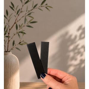May include: Two rectangular black strips with a textured surface, labeled as "Unsere Darks" and "SCHWARZ - GRAHIT (SCHIMMERND)". The strips have a matte finish and are positioned vertically, overlapping slightly to form a V-shape against a neutral background.