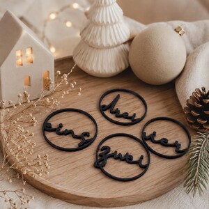 Könnte beinhalten: Vier kreisförmige schwarze Ornamente mit den deutschen Wörtern eins, zwei, drei und vier in Schreibschrift. Jedes Ornament besteht aus einem dünnen schwarzen Ring, der den handgeschriebenen Text einrahmt und als Dekoration dient.