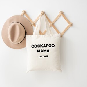 May include: A cream-coloured tote bag with the text "COCKAPOO MAMA EST 2021" in black. A beige hat with a black band hangs to the left. The bag is suspended from a wooden wall rack.