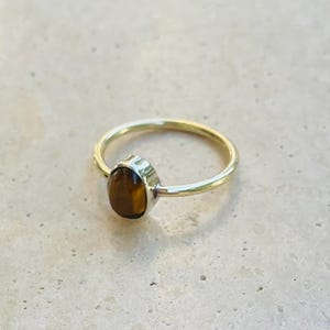 Pu&ograve; includere: Un anello color oro con una pietra marrone di forma ovale. La fascia dell'anello &egrave; sottile e liscia, e la pietra &egrave; incastonata in una lunetta. L'anello &egrave; esposto su una superficie chiara.