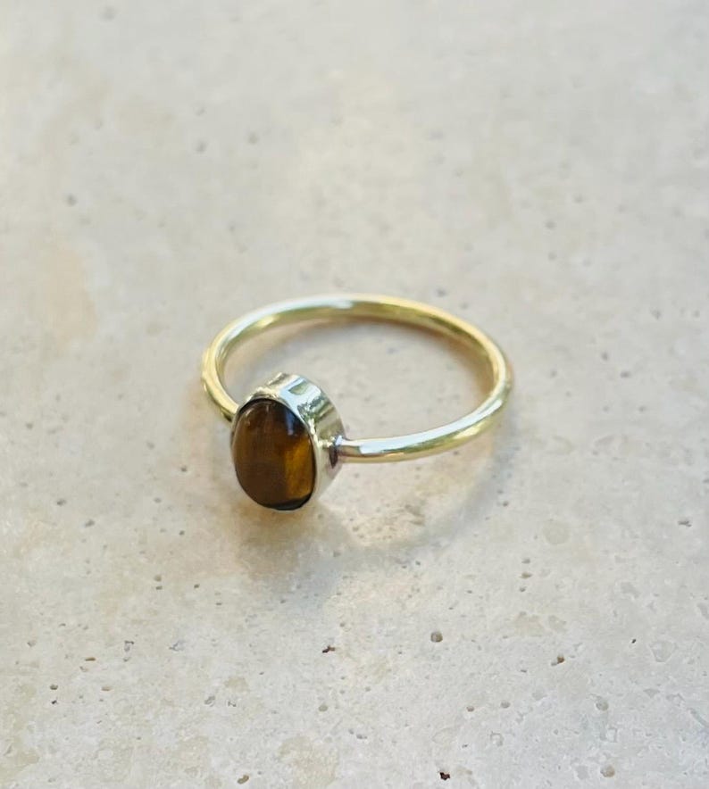 Pu&ograve; includere: Un anello color oro con una pietra marrone di forma ovale. La fascia dell'anello &egrave; sottile e liscia, e la pietra &egrave; incastonata in una lunetta. L'anello &egrave; esposto su una superficie chiara.