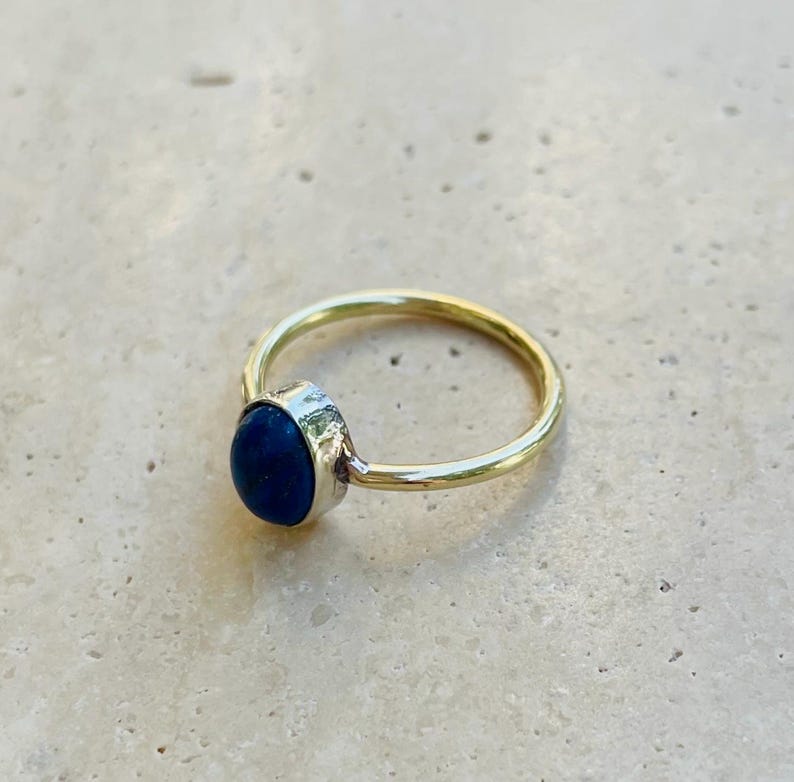 Pu&ograve; includere: Un anello d'oro con una pietra preziosa ovale blu scuro. La pietra &egrave; incastonata in una lunetta d'argento e la fascia &egrave; in oro lucido semplice. L'anello &egrave; mostrato su una superficie chiara e strutturata.
