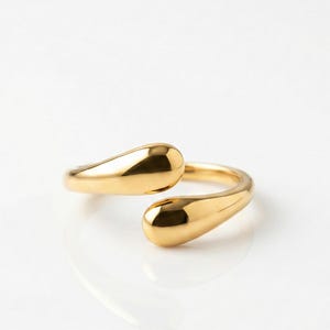 Könnte beinhalten: Ein goldfarbener Ring mit einem einzigartigen Design. Der Ring hat zwei tropfenförmige Enden, die sich umeinander krümmen. Der Ring ist vor einem weißen Hintergrund platziert, der seine polierte, reflektierende Oberfläche hervorhebt.