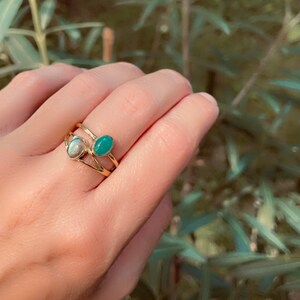Pu&ograve; includere: Una mano che indossa tre anelli d'oro. Un anello presenta una pietra ovale azzurra e un altro ha una pietra ovale verde. Il terzo anello &egrave; un design a doppia fascia. Lo sfondo &egrave; un fogliame verde sfocato.