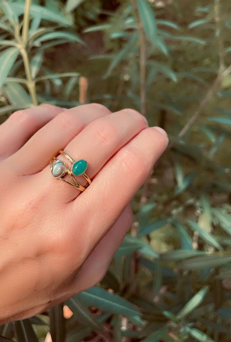 Pu&ograve; includere: Una mano che indossa tre anelli d'oro. Un anello presenta una pietra ovale azzurra e un altro ha una pietra ovale verde. Il terzo anello &egrave; un design a doppia fascia. Lo sfondo &egrave; un fogliame verde sfocato.