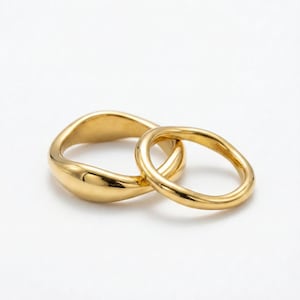Könnte beinhalten: Zwei goldene Ringe. Ein Ring ist ein glattes, breites Band mit organischer Form. Der zweite Ring ist ein dünneres, kreisförmiges Band, das sich mit dem ersten verschränkt. Beide Ringe haben eine polierte, reflektierende Oberfläche.