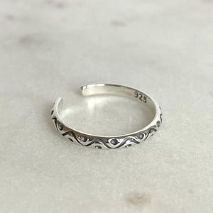 May include: A silver toe ring featuring a wave and dot design. The open ring is marked with "925" on the inside. The ring is adjustable and made of a shiny metal. The ring's design is a series of repeating patterns.