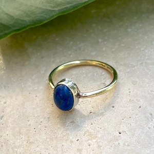 Pu&ograve; includere: Un anello d'oro con una vibrante gemma ovale blu. La fascia dell'anello &egrave; in oro lucido e la gemma &egrave; incastonata in una lunetta color argento. L'anello &egrave; esposto su una superficie chiara.