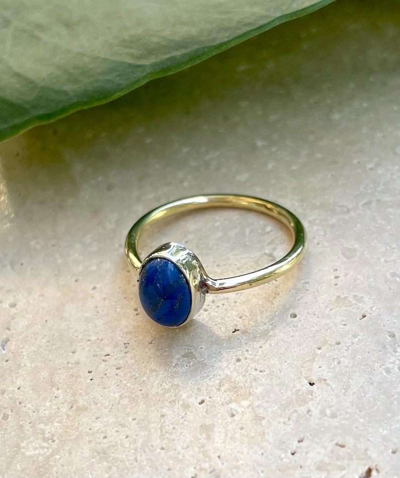 Pu&ograve; includere: Un anello d'oro con una vibrante gemma ovale blu. La fascia dell'anello &egrave; in oro lucido e la gemma &egrave; incastonata in una lunetta color argento. L'anello &egrave; esposto su una superficie chiara.