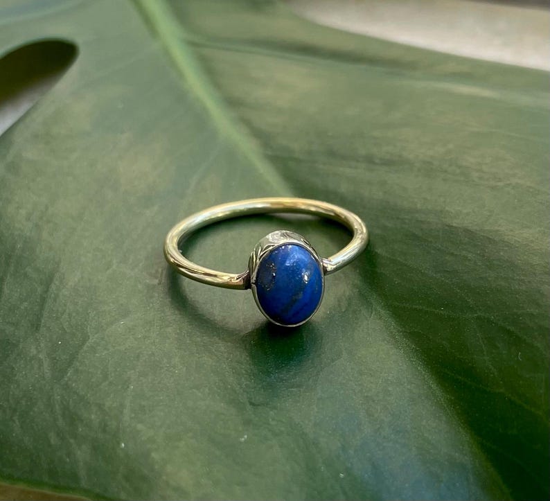 Pu&ograve; includere: Un anello d'oro con una vibrante gemma ovale blu. L'anello &egrave; posto su uno sfondo verde, evidenziando il contrasto tra la fascia d'oro e la pietra blu. Il design dell'anello &egrave; semplice ma elegante, adatto all'uso quotidiano.