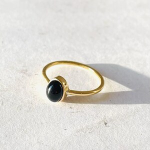 Könnte beinhalten: Ein goldfarbener Ring mit einem schwarzen, ovalförmigen Stein. Der Ring hat ein schmales Band und ein schlichtes Design. Der Stein ist in einer Zargenfassung gefasst, und der Ring wird auf einer weißen Oberfläche präsentiert.