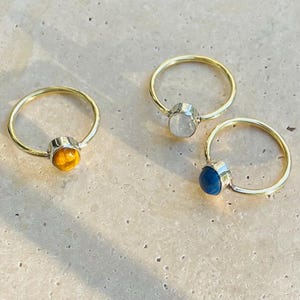 Pu&ograve; includere: Tre anelli con fasce dorate e montature argentate. Ogni anello presenta una gemma diversa: un occhio di tigre giallo-arancio, una pietra di luna bianca e una pietra blu scuro. Gli anelli sono esposti su una superficie chiara.