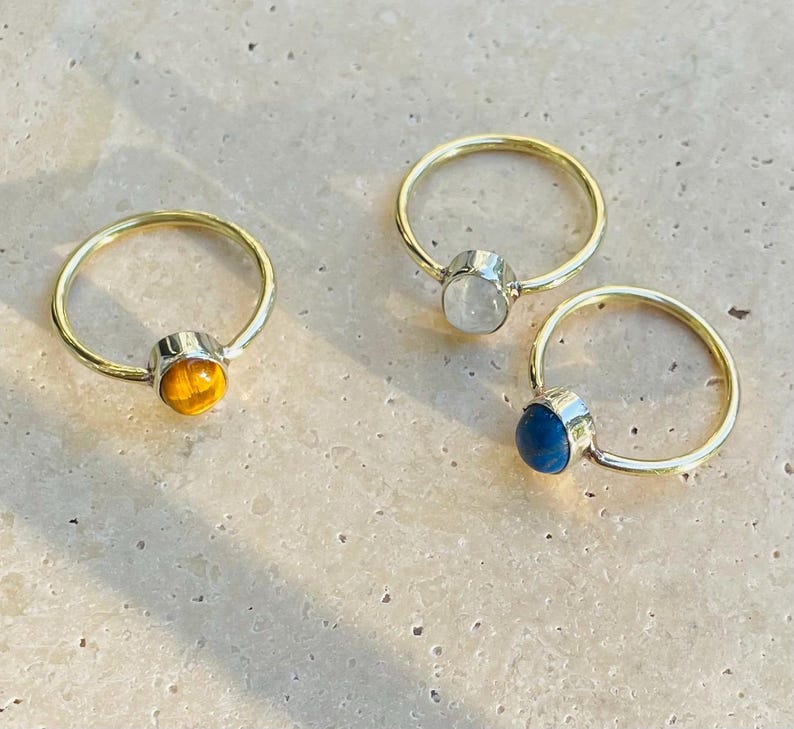 Pu&ograve; includere: Tre anelli con fasce dorate e montature argentate. Ogni anello presenta una gemma diversa: un occhio di tigre giallo-arancio, una pietra di luna bianca e una pietra blu scuro. Gli anelli sono esposti su una superficie chiara.