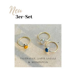 Pu&ograve; includere: Tre anelli dorati con pietre preziose. Un anello ha una pietra giallo-marrone, un altro una pietra trasparente e il terzo una pietra blu. Il testo nell'immagine recita "New 3er-Set" e "TIGERAUGE, LAPIS LAZULI & MONDSTEIN".