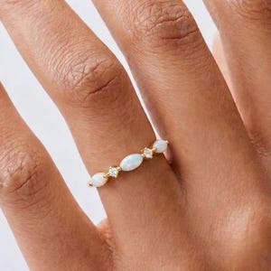 Könnte beinhalten: Ein goldener Ring mit ovalen Opalen und kleinen Diamanten. Die weißen Opale schimmern leicht, eingefasst in einem schmalen Goldband. Der Ring wird an einem Finger getragen. Ein elegantes Schmuckstück, das durch die Diamanten funkelt.