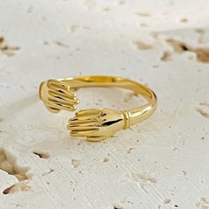 Könnte beinhalten: Ein goldfarbener Ring mit dem Design von zwei sich umarmenden Händen. Der Ring besteht aus poliertem Metall und liegt auf einer strukturierten, cremefarbenen Oberfläche. Die Hände sind mit einzelnen Fingern detailliert.