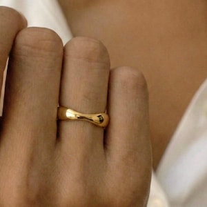 Könnte beinhalten: Ein goldfarbener Ring mit einem einzigartigen, geschwungenen Design. Der Ring ist glatt und glänzend, mit einer leicht sanduhrförmigen Form. Der Ring wird an einem Finger getragen, der Hintergrund ist unscharf.