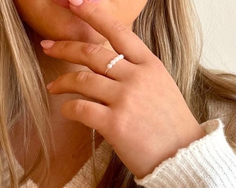 May include: A delicate gold ring with a row of small white pearls. The ring is worn on a person's finger.