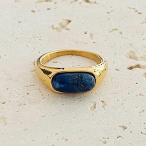 Könnte beinhalten: Ein goldfarbener Ring mit einem rechteckigen, tiefblauen Stein. Der Ring hat einen glatten, polierten Ring und eine Lünettenfassung. Der Stein ist etwa 1,5 cm lang. Es ist ein Schmuckstück.